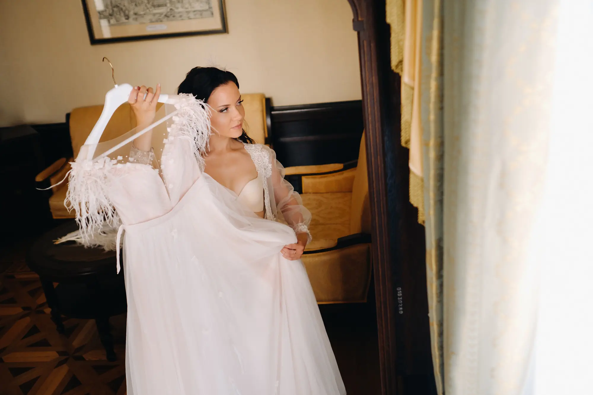 a bride dressed in a boudoir transparent dress and underwear holds her wedding dress in her hands in