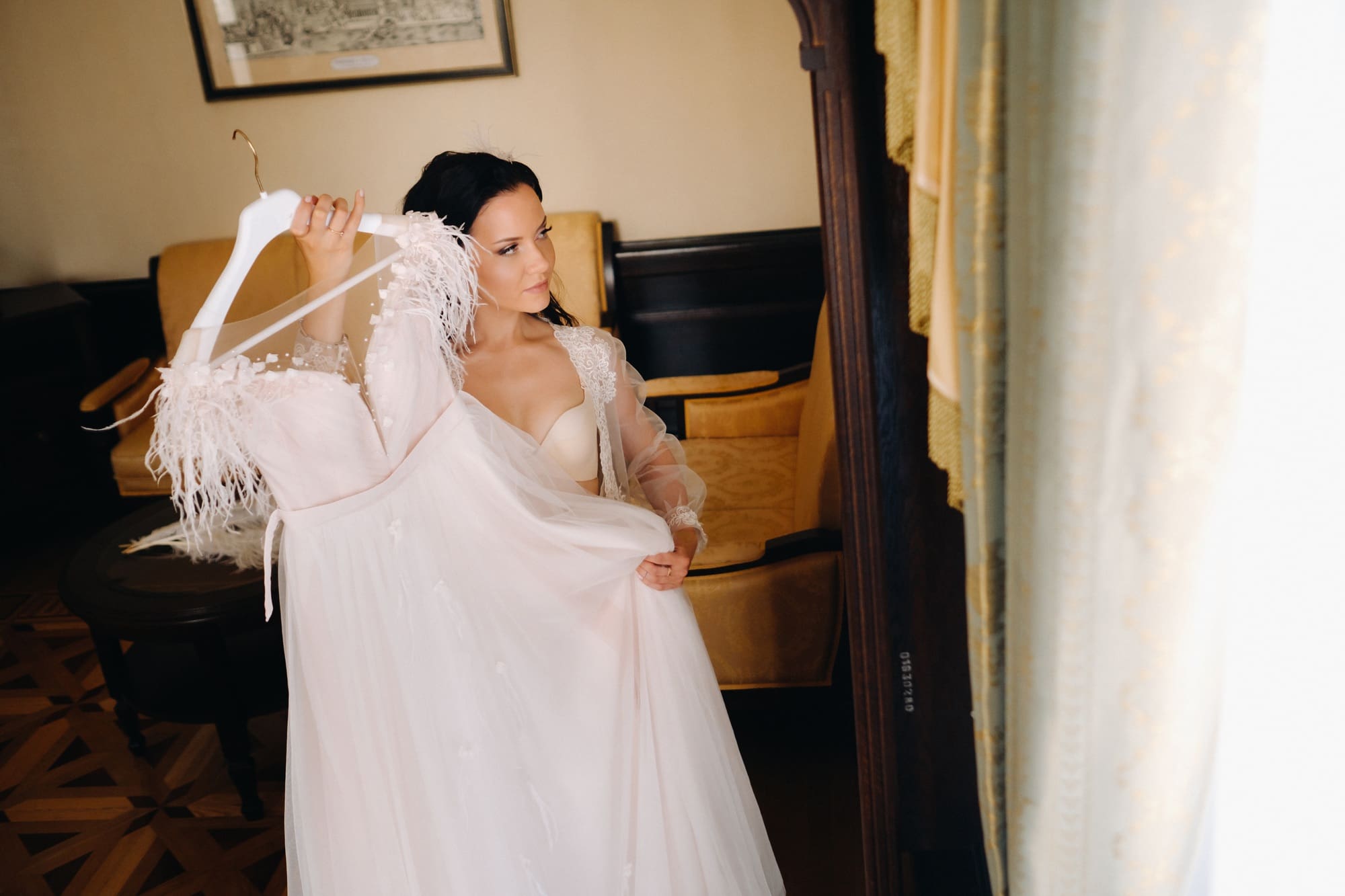 a bride dressed in a boudoir transparent dress and underwear holds her wedding dress in her hands in